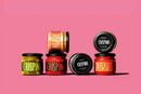 Five jars of Taycte CRISPINO sauces in an array of colors are arranged on a vivid pink background, some standing upright and others lying on their sides, showcasing eye-catching black, green, and red labels. This vibrant assortment includes vegan and gluten-free choices such as Taycte Crispino Bruschetta Pomodori Secchi — Calabrian Sun-Dried Tomato Spread, making the collection as delicious as it is visually striking.