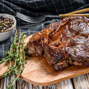 A juicy, grilled steak rests on a wooden board, garnished with fresh thyme and SALTOPIA Infused Sea Salts' SCARBOROUGH FAIR Rosemary Salt. Nearby are mixed peppercorns, a fork, and a checkered napkin to complete the savory scene.