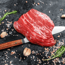 The Dude Food Essentials Meat Box by Dude Food is presented on a dark surface, featuring raw flank steak atop a wooden-handled knife and surrounded by rosemary sprigs, garlic cloves, and coarse pink salt—an enticing scene that inspires meat lovers to create mouthwatering flat iron steak dishes.