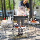 A man in an apron grills food on a DirectDeals Foldable Outdoor BBQ Table Grilling Stand, surrounded by drinks, food, and utensils, with trees and a camper in the background.