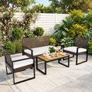 The DirectDeals 4-Piece Patio Conversation Set with Acacia Coffee Table features white cushions and sits on a rug in a lush garden, with a tea set, snacks, flowers, bushes, and a white fence in the background.