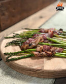 Grilled asparagus wrapped in crispy bacon is artfully arranged on a round wooden board, demonstrating the mouthwatering results made possible with the WPPO LLC Direct Professional Lava Digital Controlled Wood-Fired Oven With Convection Fan. The WPPO Get Fired Up logo appears in the top right corner.