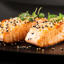 Two pieces of cooked salmon on a dark plate, topped with white sesame seeds and The Spice Lab Black Sesame Seeds for a delightful nutty flavor, and garnished with fresh green herbs.