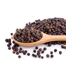 A wooden spoon brimming with whole Tellicherry Peppercorns lies on a white surface, surrounded by scattered peppercorns—perfect for filling your The Spice Lab Premium Black Pepper Grinder by The Spice Lab, so you can enjoy the bold flavor of freshly ground pepper.