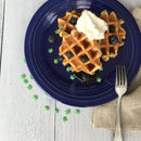 Two waffles topped with blueberries, syrup, and whipped cream sit on a blue plate, with a fork on a beige napkin beside. The table is decorated with The Spice Lab Hemp Leaf Sprinkles CBD & THC Free by The Spice Lab—small green edible party decorations that are completely free of CBD and THC.