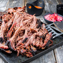 Shredded smoked brisket on a tray with a spatula, served atop a rustic wooden table, accompanied by pickled red onions and a cup of orange sauce in the background—perfectly seasoned with The Spice Lab Cooking Essentials Seasoning Set from The Spice Lab to enhance every savory bite.