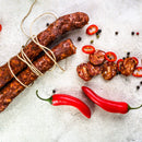 Three tied dried sausages, sliced sausage pieces with red chili pepper slices, two whole red chili peppers, and scattered peppercorns on a light surface—all perfectly accented with a hint of The Spice Lab Organic Spanish Sweet Paprika to enhance the flavor profile.