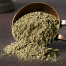 A copper measuring cup spills a mound of fine, greenish-brown powder onto a dark surface, showcasing The Spice Lab Organic Rubbed Sage—an aromatic seasoning perfect for stuffing and poultry dishes.