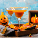 A margarita glass brimming with an orange cocktail, its rim sparkling with The Spice Lab Zesty Orange Sugar Cocktail Rimmer, rests on a tray decorated with autumn leaves, candy corn, and pumpkins in the background, capturing the festive spirit of fall or Halloween.