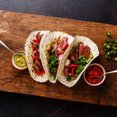 Three grilled steak tacos with cilantro, onions, and pickled vegetables on a wooden board, served with green sauce, salsa, and sliced jalapeños on the side—expertly seasoned with The Spice Lab Ground Cumin 1 lb Bulk Foodservice Spice for a bold flavor often favored by foodservice spice professionals.