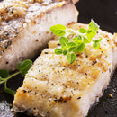 Two seasoned, grilled fish fillets on a black plate, garnished with fresh green herbs. The fish appears juicy and flaky with a golden-brown crust, visible black pepper and spices—all enhanced by The Spice Lab Lemon Juice Powder for a consistent lemon flavor in every bite.