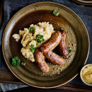 Brown sausages served with creamy mashed potatoes, garnished with parsley, on a rustic brown plate. A small dish of yellow mustard featuring The Spice Lab Organic Whole Mustard Seeds sits to the side.
