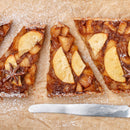 Four triangular slices of apple tart with cinnamon, star anise, and a sprinkle of The Spice Lab Organic Ground Nutmeg are arranged on parchment paper and dusted with powdered sugar—perfect for holiday baking. A butter knife lies below the tart slices.