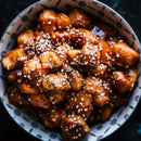 A bowl of glazed chicken pieces topped with The Spice Lab Organic Hulled Sesame Seeds, served in a patterned bowl on a dark background.