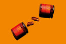 Two glass jars labeled CRISPY lie on their sides on an orange background, with Taycte Famiglia Crispino Sun-Dried Tomato Antipasto — Italian Marinated in Olive Oil spilling between them.
