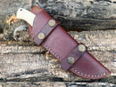 The Texan Knives Custom Handmade Damascus Bushcraft Knife features a white camel bone handle and comes in a brown leather sheath with metal accents and white stitching, shown resting on rugged wooden logs.