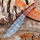 The Texan Knives Custom Handmade 5-Inch Damascus Skinning Knife with a patterned blade and rosewood handle rests on rustic wood, with its brown leather sheath displayed above it.