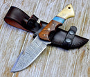 The Texan Knives Premium Damascus Guthook Skinning Knife features a 10” patterned blade and a rosewood & bone handle, resting on its brown leather sheath atop a wooden surface.