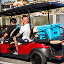 Three people ride a red golf cart near a waterfront. A large blue Bob The Cooler Co. 50 QT/47.3L Hard Cooler - El Amigo, featuring a removable dry basket, sits on the back. Houses and palm trees are visible across the water under a sunny sky.