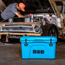 A man works under the hood of a rusty classic car in a garage. In the foreground, a bright blue Bob The Cooler Co. 50 QT/47.3L Hard Cooler - El Amigo by Bob - The Cooler Co., featuring a removable dry basket, sits on the concrete floor.