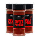 Three jars of The Spice Lab Sweet Paprika Powder are lined up in a row. Each clear plastic container displays the bright red hue of the paprika powder inside, featuring a black label with bold red lettering that reads “SWEET PAPRIKA.”.