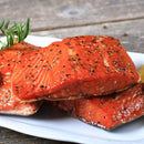 Three pieces of cooked salmon fillet seasoned with freshly ground The Spice Lab Premium Tellicherry Peppercorns from the convenient grinder sit on a white plate with a sprig of rosemary, atop a rustic wooden table.