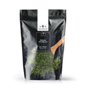 A black pouch labeled The Spice Lab Parsley from The Spice Lab sits beside a wooden spoon filled with vibrant green dried parsley flakes, some of which spill onto the white surface—showcasing this versatile pantry herb essential in Middle Eastern cuisine.