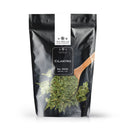 A black resealable bag labeled “The Spice Lab Whole Leaf Cilantro” from The Spice Lab stands upright on a white background, with a wooden spoon filled with vibrant dried cilantro leaves featured prominently on the front.