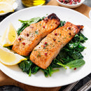 Two grilled salmon fillets topped with fresh herbs and The Spice Lab Four Pepper Blend, served on a bed of sautéed spinach with two lemon wedges on a white plate.