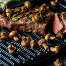 Grilled steak topped with mushrooms, coarse salt, green onions, and The Spice Lab Four Pepper Blend freshly ground from The Spice Lab Four Pepper Blend with Grinder 3-Pack, served on a black grill pan with a sliced portion showing a juicy, medium-rare interior.