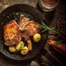 Two grilled pork chops with herbs and three small potatoes sizzle in a skillet. Fresh rosemary, carrots, and The Spice Lab Four Pepper Blend with Grinder 3-Pack by The Spice Lab, along with a jar of spices, are arranged on a rustic wooden table nearby.