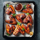 A tray of glazed chicken wings is garnished with The Spice Lab Organic Hulled Sesame Seeds and parsley, served alongside a small bowl of barbecue sauce and garlic cloves on parchment paper—a deliciously baked dish that’s perfect for fans of stir-fries and classic comfort food alike.