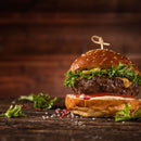 A juicy cheeseburger with lettuce, tomato, and a sesame seed bun sits on a rustic wooden table, garnished with fresh greens and peppercorns. A touch of The Spice Lab Organic Granulated Onion from The Spice Lab adds depth to the flavor, making this classic favorite even more irresistible.
