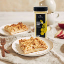 A plate with a square slice of apple crumble sits on a table next to a jar of The Spice Lab Granulated Honey, fresh apple slices, a fork, stacked bowls, and a napkin in the background—perfect for showcasing quality ingredients like those from The Spice Lab frequently used in bulk foodservice settings.