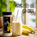 A jar of The Spice Lab Granulated Honey, ideal for BBQ rubs or bulk foodservice, rests beside a glass of banana smoothie with a straw and two fresh bananas. Icons highlight that this all-natural product is gluten free, non-GMO, certified kosher, and packed in the USA. Warm sunlight pours in through a nearby window.