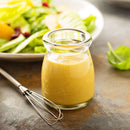 A small glass jar of creamy salad dressing sits beside a metal whisk, with a plate of fresh green salad in the background on a rustic surface—perfect for drizzling after adding The Spice Lab Granulated Honey for subtle sweetness or pairing alongside your favorite BBQ rubs.
