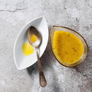 Two leaf-shaped dishes rest on a textured surface: one holds a spoon with a small amount of yellow vinaigrette, while the other is filled with more vinaigrette speckled with black pepper. The flavors—reminiscent of classic BBQ rubs—are enhanced by the subtle sweetness of The Spice Lab Granulated Honey from The Spice Lab.