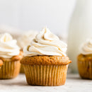 A close-up of a cupcake featuring light brown cake, delicately sweetened with The Spice Lab Maple Sugar Powder, and topped with a generous swirl of creamy white frosting, while more cupcakes from the same batch are softly blurred in the background.