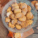 A glass plate filled with assorted autumn-themed cookies shaped like leaves and acorns, dusted with powdered sugar and a hint of The Spice Lab Maple Sugar Powder by The Spice Lab, sits on a wooden board with decorative fall leaves nearby.