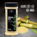 A container of The Spice Lab Turbinado Sugar rests on a dark surface, accompanied by sugar cane stalks and a small pile of loose sugar. The label emphasizes that it is all-natural, gluten-free, non-GMO, and packed in the USA. This less processed sugar from The Spice Lab is perfect for sweetening your favorite recipes while preserving a natural touch.