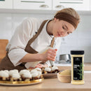 A woman in a chef’s uniform decorates cupcakes with icing in a kitchen. On the counter in the foreground are a bowl and a container of The Spice Lab Turbinado Sugar, an unrefined sweetener from The Spice Lab that’s perfect for adding natural sweetness.