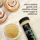 A jar of The Spice Lab Turbinado Sugar sits beside a bowl filled with this less processed sugar and three cinnamon rolls on a cooling rack. Text reads: Use The Spice Lab Turbinado Sugar in baked goods and pastries where white sugar is called for—perfect for sweetening your favorite treats.