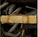 A Texan Knives 13" Handmade Damascus Steel Chef Knife with epoxy resin handle and leather sheath sits outdoors on stacked firewood, ready to protect your premium kitchen knife set or prized chef knife.