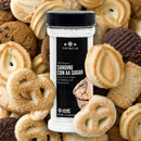A jar of Sanding Con AA Sugar from The Spice Lab is surrounded by assorted butter cookies, including pretzel and swirl shapes—perfect for decorative baking or as sprinkles on cupcakes.