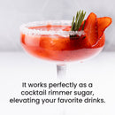 A cocktail with a sugar-rimmed glass, garnished with strawberry slices and rosemary, sits on a white surface with text beneath describing The Spice Lab’s Sanding Con AA Sugar—vibrant sanding sugar perfect for rimming cocktails or adding a festive touch to decorative baking and cupcake sprinkles.