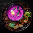 A bowl of vibrant pink soup with half a boiled egg, garnished with The Spice Lab Organic Whole Dill Weed, served on a plate with roasted potatoes and fresh greens on the side, all set against a dark background.
