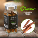 A glass jar labeled The Spice Lab Organic Cinnamon Sticks sits on a wooden surface, featuring a silver lid and filled with organic cinnamon sticks from The Spice Lab. A close-up circle highlights loose cinnamon sticks beside the jar, perfect for adding warm spice to your favorite chai or mulled wine.