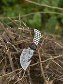 The Texan Knives Custom Handmade Damascus Steel Guthook Skinning Knife, 7.5” with a rosewood & resin handle and included leather sheath, rests on dry branches with softly blurred green foliage in the background.