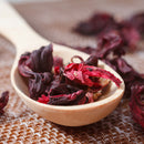 A wooden spoon brimming with The Spice Lab Whole Hibiscus Flowers, featuring whole hibiscus petals as a premium botanical ingredient from The Spice Lab, rests on a woven brown surface, with extra dried petals scattered in the background.