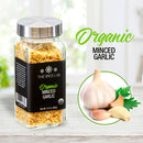 A jar labeled The Spice Lab Organic Minced Garlic sits beside a plate with fresh garlic bulbs and cloves, and parsley—perfect for sprinkling over avocado toast or using as a substitute for dried garlic. The label features black with white and yellow text. The background is bright and softly blurred.